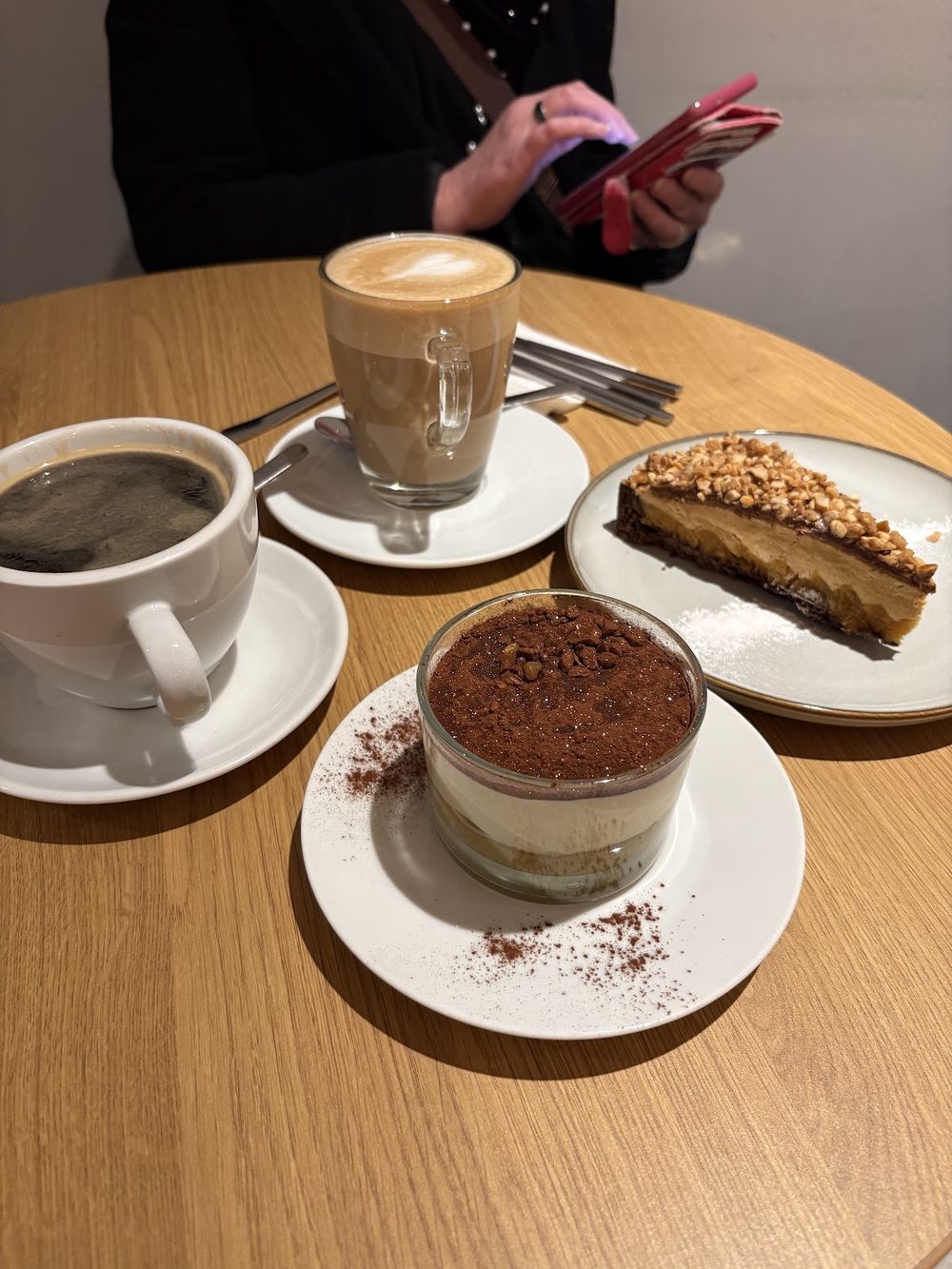 A table set with a latte macchiato, an americano, tiramisu in a glass and a caramel tart slice, someone sitting further back in the room
