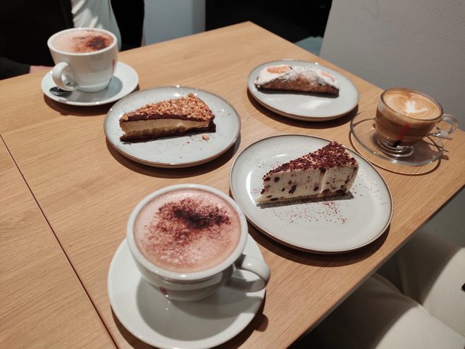 A shared wooden table with two cakes, a cheesecake slice, a cappuccino and a latte macchiato