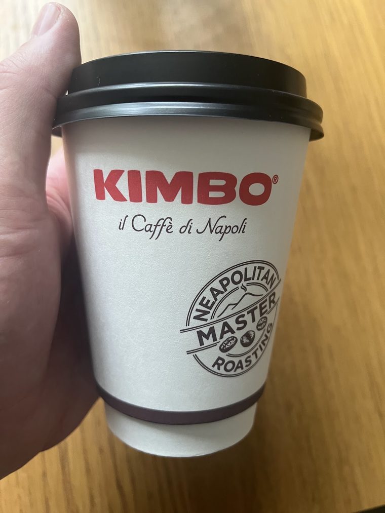 A takeaway coffee cup held against a wooden background, cropped tight to the hand and cup so the burned-in wording is out of frame